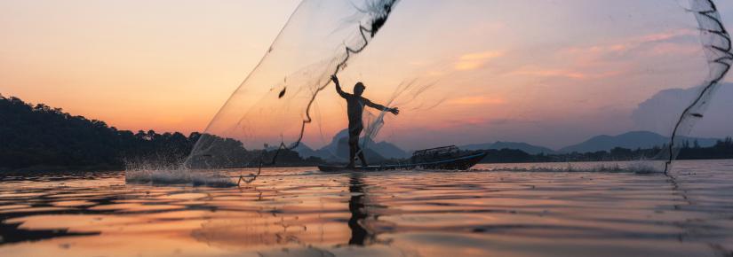 Fishermen throwing fishing net during twilight on wooden boat at the lake.