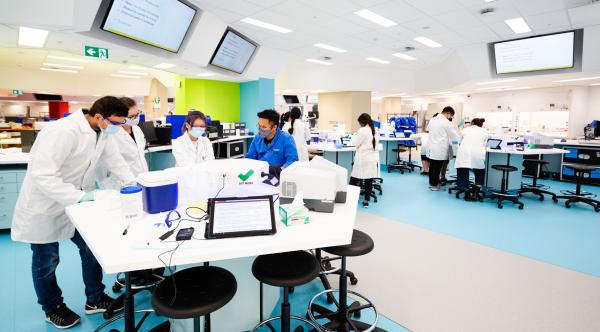 Students in white lab coats work around hexagonal desks filled with science equipment. Walls and floors are coloured blue, green and beige.