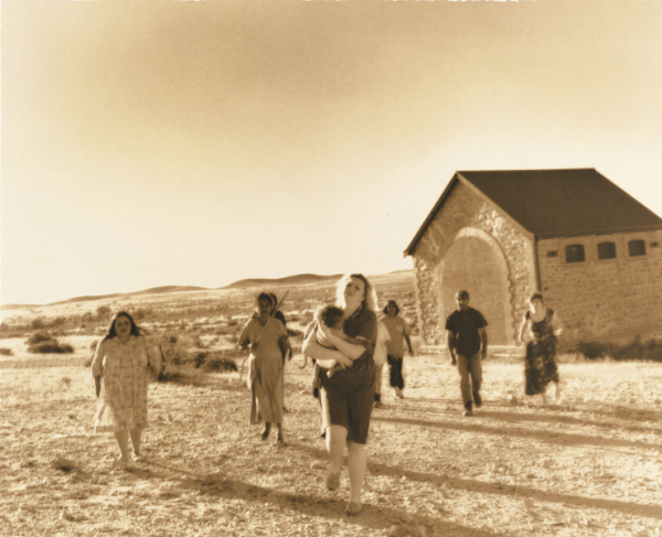 A sepia toned image of a woman holding a baby running with people behind her. A church stands in the background. 