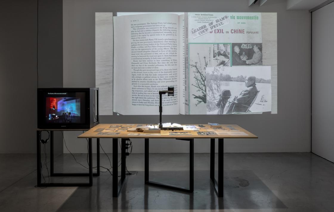 a sculptural installation by Prima Jalichandra-Sakuntabhai, which comprises a large timber table with carefully placed archival materials and a digital document camera which is projecting a collage of these materials onto the wall beside the table. There is a smaller table of the same height and materials which holds a heavy 1990’s-era TV monitor with headphones on a hook below the screen