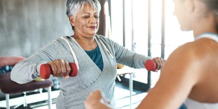 Senior female client and female trainer using dumbbells. Client has short grey haird and is wearing a grey sweatshirt. The trainer is wearing a sports tank top and has her hair in a pony tail..