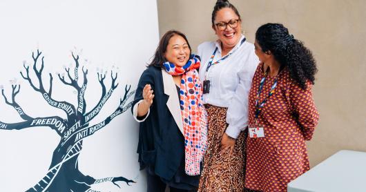 Three women laughing and smiling next to the anti-racism tree artwork