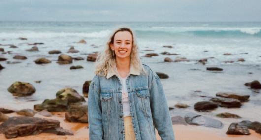 Photo of girl standing on the beach with the ocean in the background. She is smiling at the camera. She has blonde hair and is wearing a denim jacket.