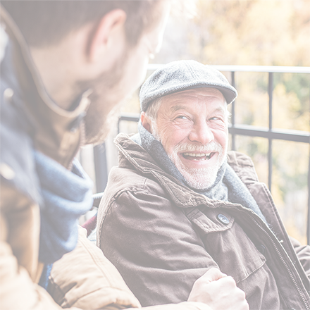 Senior father in wheelchair and young son on a walk. The father has a beard and is wearing a flat cap, coat and scarf. The son also has a beard and is wearing a coat and scarf. They are looking at each other and smiling.  