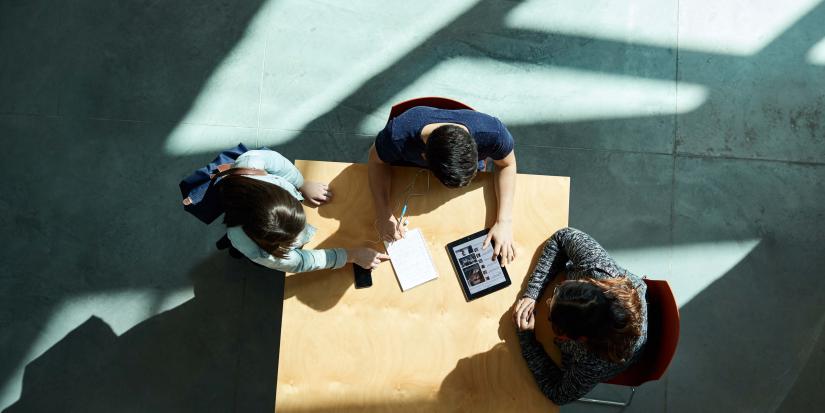 Overhead photo of three students sitting at a table with a notepad and an iPad