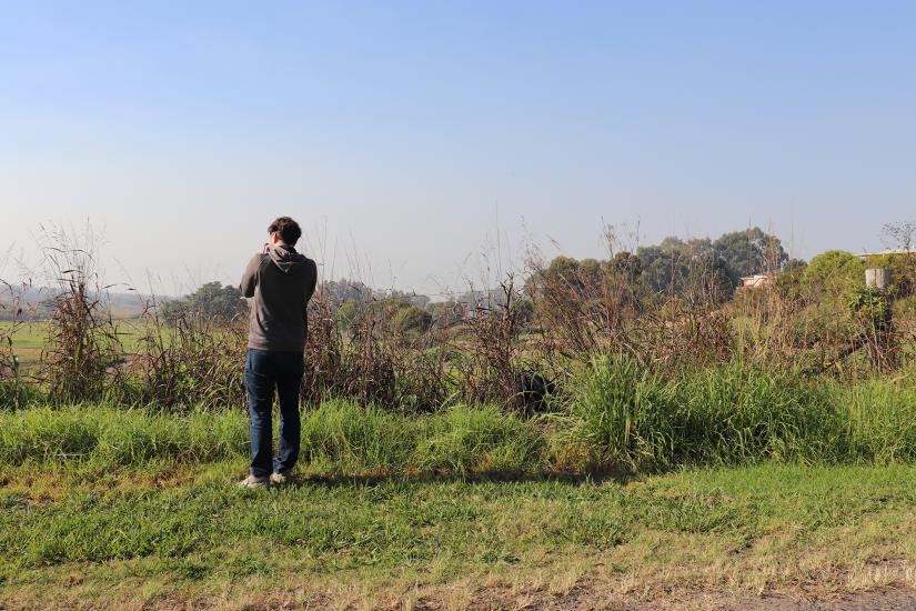 The back of a student taking a photo, standing in a grassy landscape.