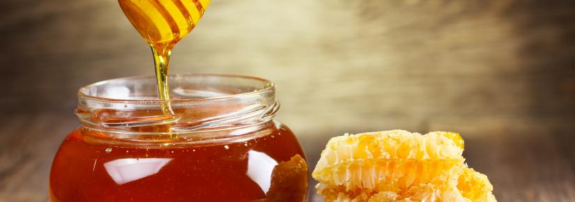 A spoon dripping with honey is held over a pot of honey next to a honeycomb.