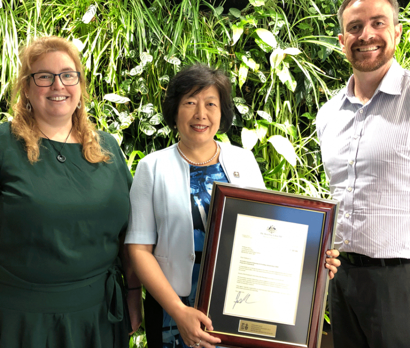 Distinguished Professor Jie Lu holding a framed certificate with Kate McGrath on her left and Attila Brungs on her right. Lush green foliage behind.