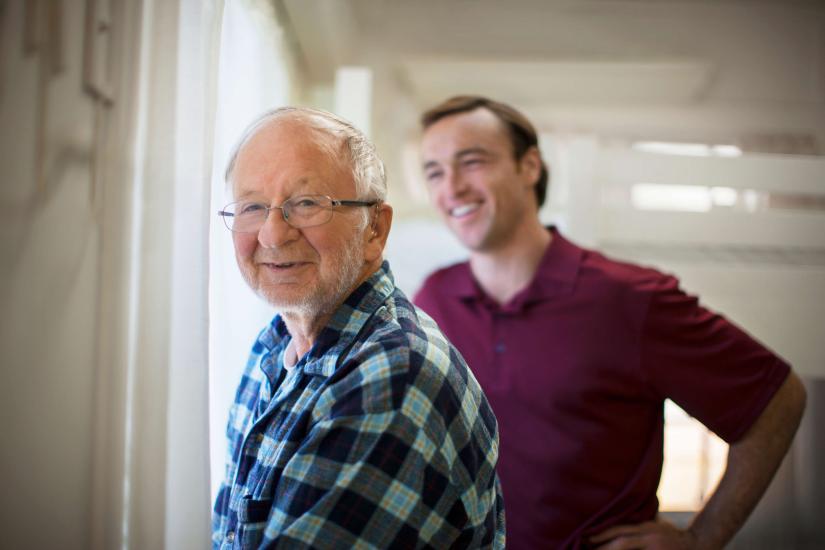 Older male patient in checked shirt smiles at camera. Male carer in red short sleeved shirt stands behind him smiling.