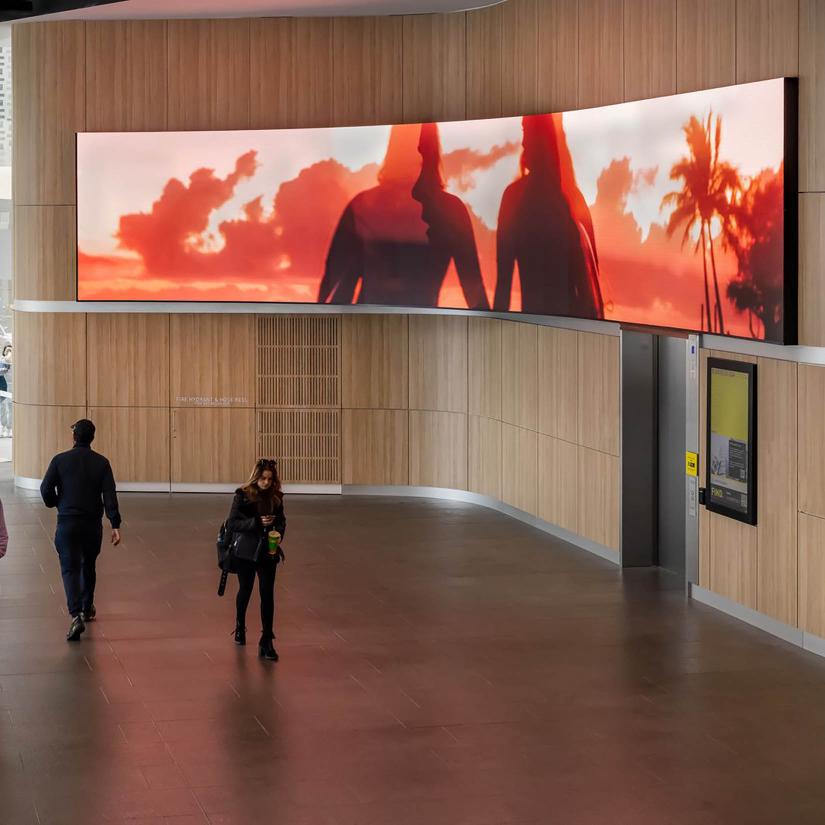 A large screen in a public space shows an image of two people holding hands against a sunset and palm tree scene. Two people in the foreground of the screen walk past. 