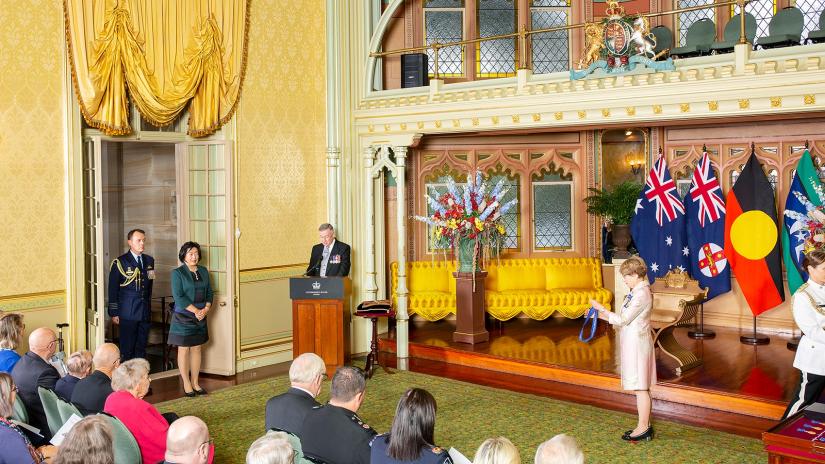 Long distance shot of Distinguished Professor Jie Lu waiting in the doorway of an official state room to receive her Order of Australia medal from the NSW Governor General, also pictured. Jie Lu is standing with her hands folded in front of her, wearing a knee length dress with thick black, emerald green and navy stripes with a matching green fitted jacket. The first few rows of the audience are visible. There are four official flags in the background, including the Australian flag and the Indigenous flag. 