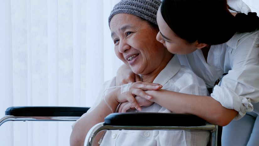 Asian lady with head scarf is sitting in a wheelchair. Her daughter is hugging her from behind. 