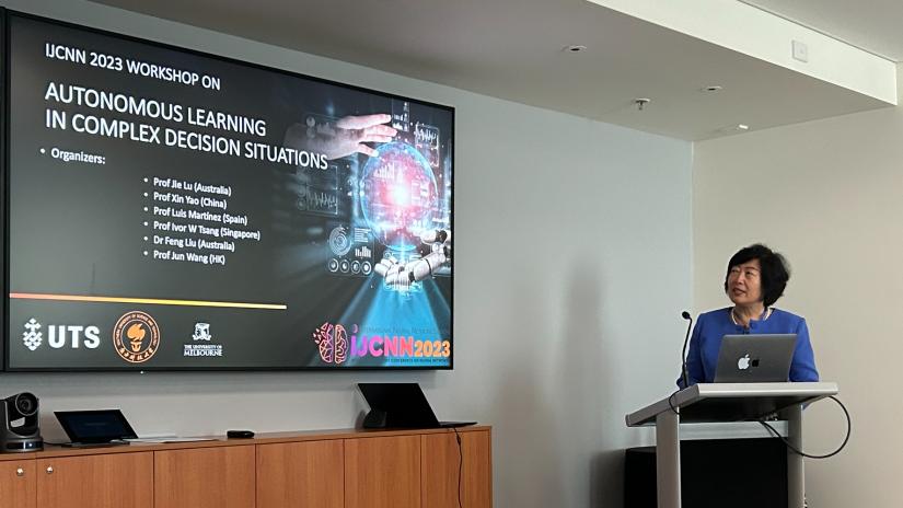 Image of Dist. Prof. Jie Lu standing behind a podium, delivering a presentation from a screen. The screen is on the title slide which reads 'Autonomous Learning in Complex Decision Situations'
