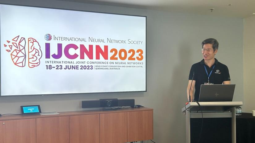 Prof. Xin Yao standing behind a podium delivering a presentation. There is a screen behind him which has the IJCNN2023 logo pictured on it. Prof Xin Yao is smiling and looking to the side. He is wearing a black t-shirt.