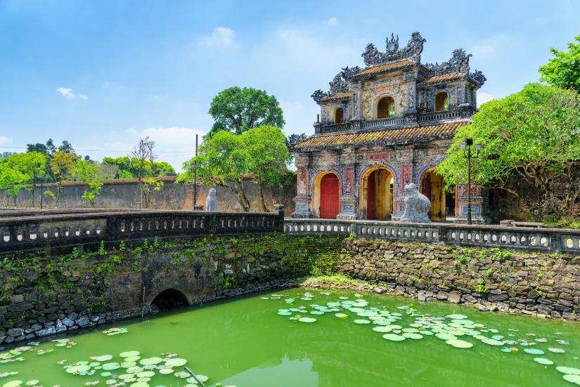 A temple in Hue, Vietnam, with a stone bridge over a green lake and ornate designs atop the entrance