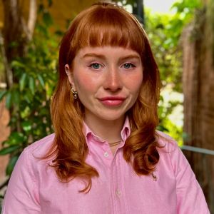 A headshot of Madeleine McWilliam from the shoulders up. Madeleine has red hair and is wearing a light pink collared shirt. The background is out of focus, featuring greenery and a brick wall.
