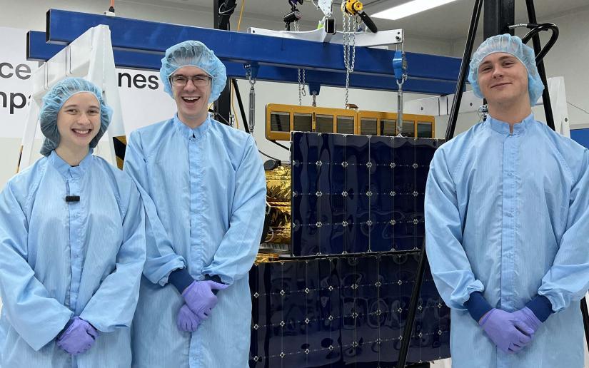 Engineering students Liv, John and Jacob standing in blue PPE next to the satellite Optimus