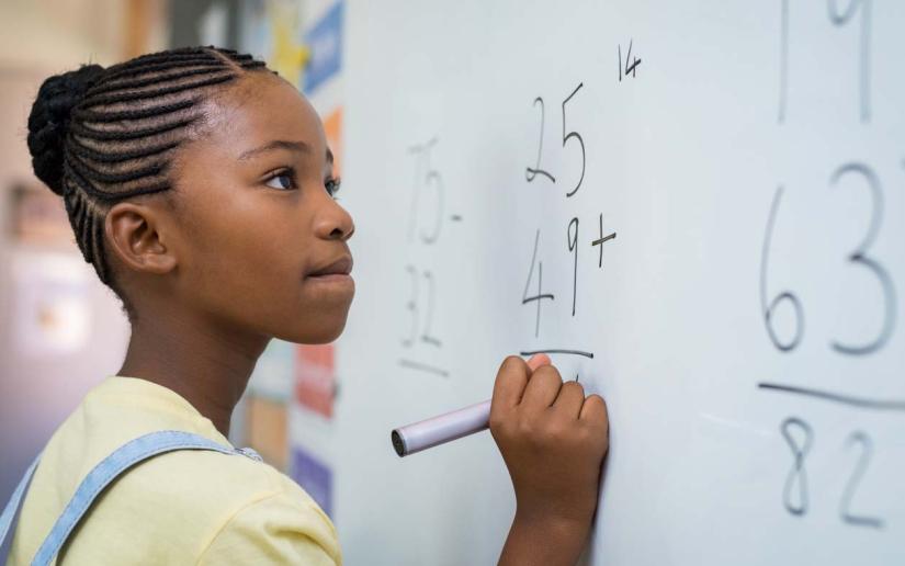 A young girl attempts a maths problem at a classroom whiteboard.
