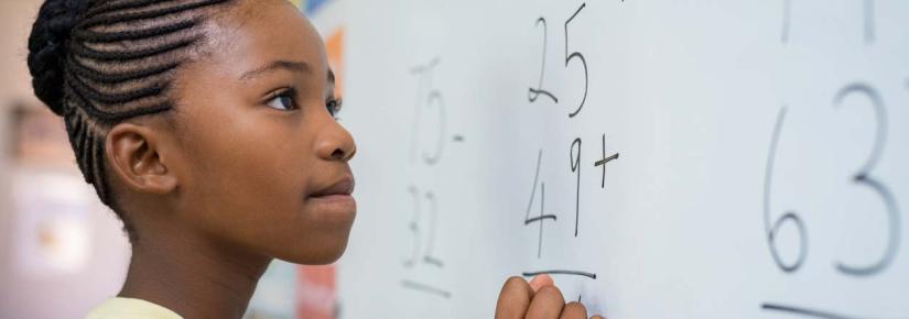 A young girl attempts a maths problem at a classroom whiteboard.