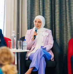 Nadine El-Kabbout is talking with a microphone on a stage at the PaCCSC & CST Annual Research Forum 2023. She is wearing mauve trousers, white shires and pale pink blazer and a white head scarf. 