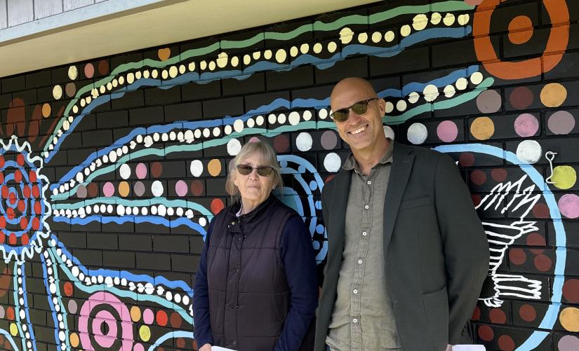 Two people smiling at the camera, an Indigenous mural behind them