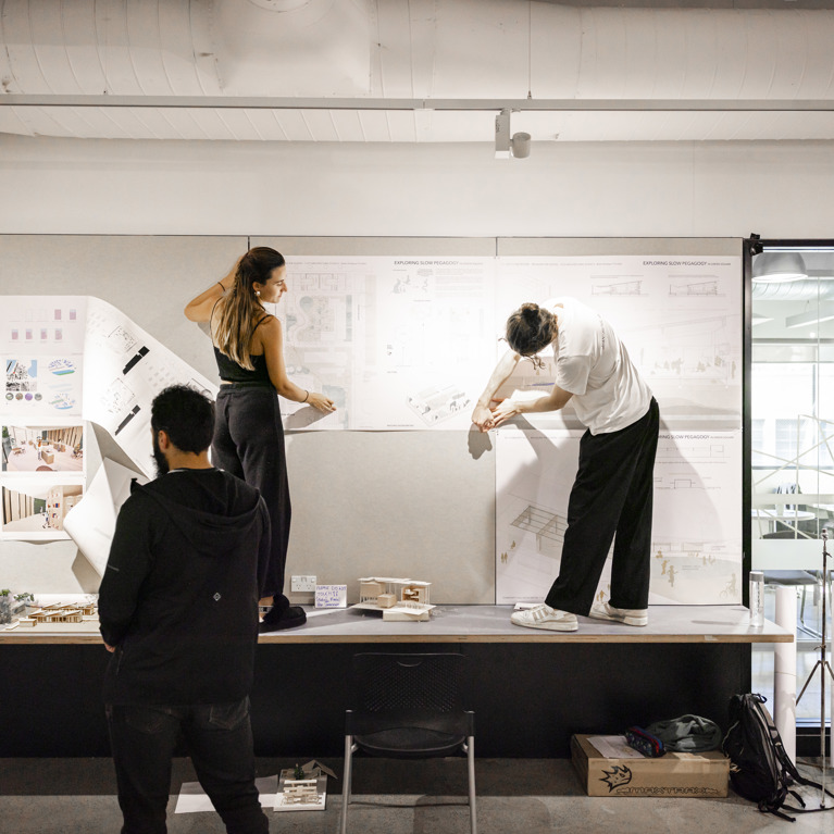 Architecture students stand on table sticking drafting drawings to the wall