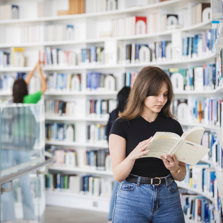 Student standing in library looking through a book.