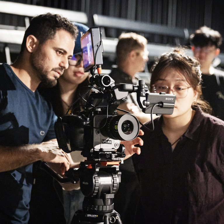 Three people, one male and two female, stand around a Sony video camera looking at what is displaying on the camera's screen. Two blurry people are in the background behind them. They are in a dark room with stage lighting on them and grandstand-style seating behind them.