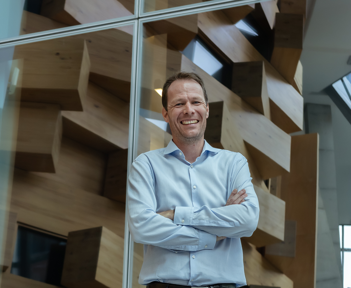 Nico Schulenkorf stands in the centre of the frame from the wasit up, the wooden Oval Classroom is behind him in the background. He has his arms crossed and his smiling to camera wearing a blue business shirt.