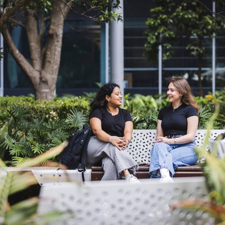 Two female students sitting on a bench talking, surrounded by green plants.