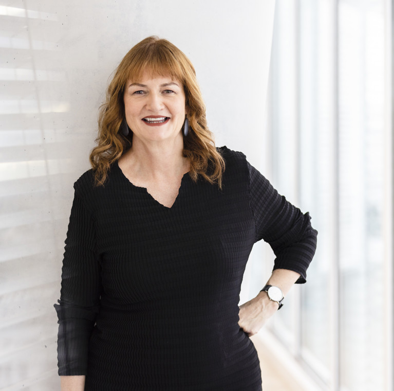 In the foreground Elizabeth Mossop smiles to camera in a black dress with one hand on her hip. In the background is a brightly lit interior of the UTS's Building 11.
