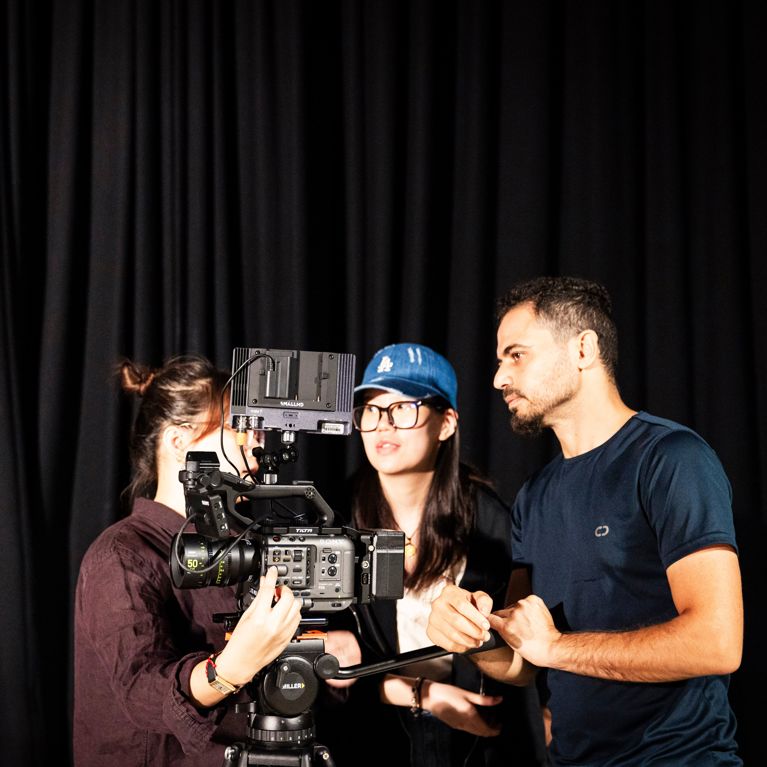 Three students standing around film camera in dark studio.