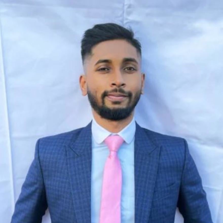 Farhan, a man stands in a blue jacket with a pink tie and short-cropped dark hair, he smiles at the camera. The background is a white tarp.