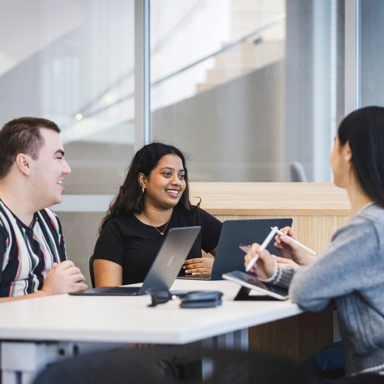 A group of students sitting around a table with laptops, talking and smiling.