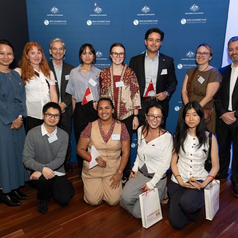 Group of scholars and representatives stand against blue media wall with Australian Government and New Colombo Plan logos. From left to right, top row: Kartika Paksi, Madeleine McWilliam, Innes Ireland, Nina Jayengrani, Amelia Wilson-Williams, Michael Farah, Alice Loda, Nick Manganas. Bottom row: Leo Chau, Kayla Baker-Peris, Emily Rutten, Lucia Guo.