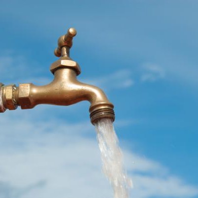 Water runs from a copper-coloured tap. The background is a blue sky.