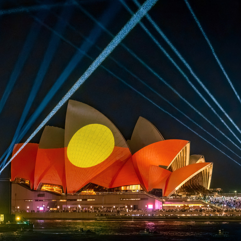 The Sydney Opera House is illuminated at night with vibrant red and yellow Indigenous flag art projections. Blue laser beams radiate into the night sky.