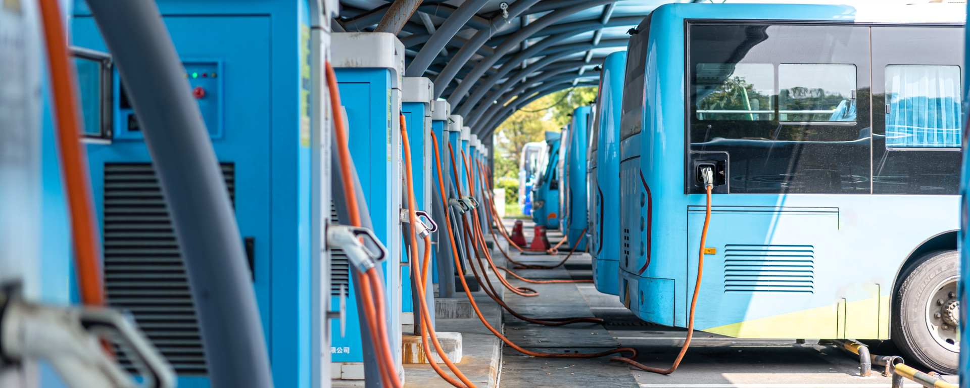 A row of electric buses is parked at a charging station, each connected to a blue charger with orange cables, under a modern canopy walkway.