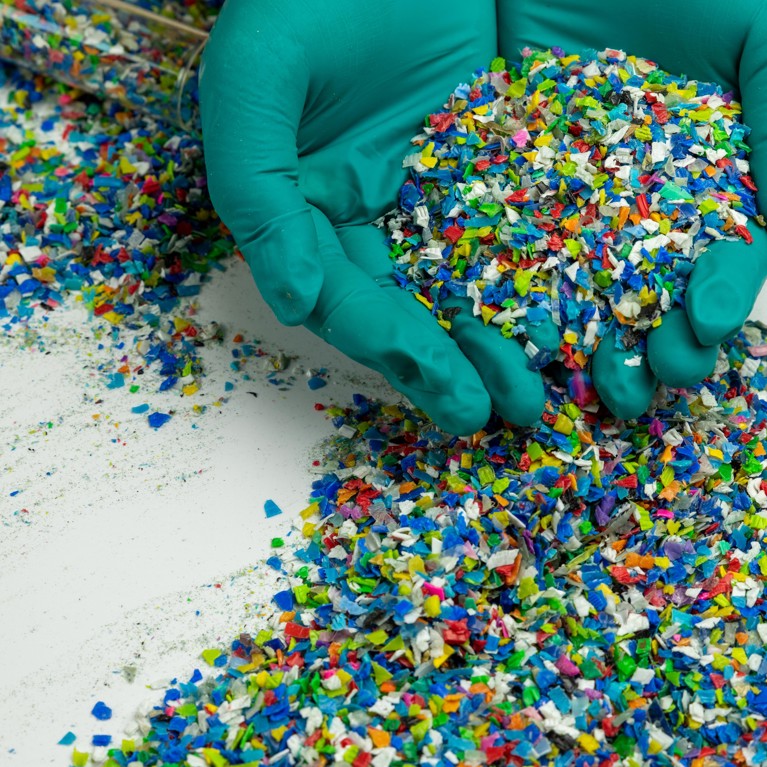 Two hands wearing green rubber gloves hold particles of coloured recycled plastic