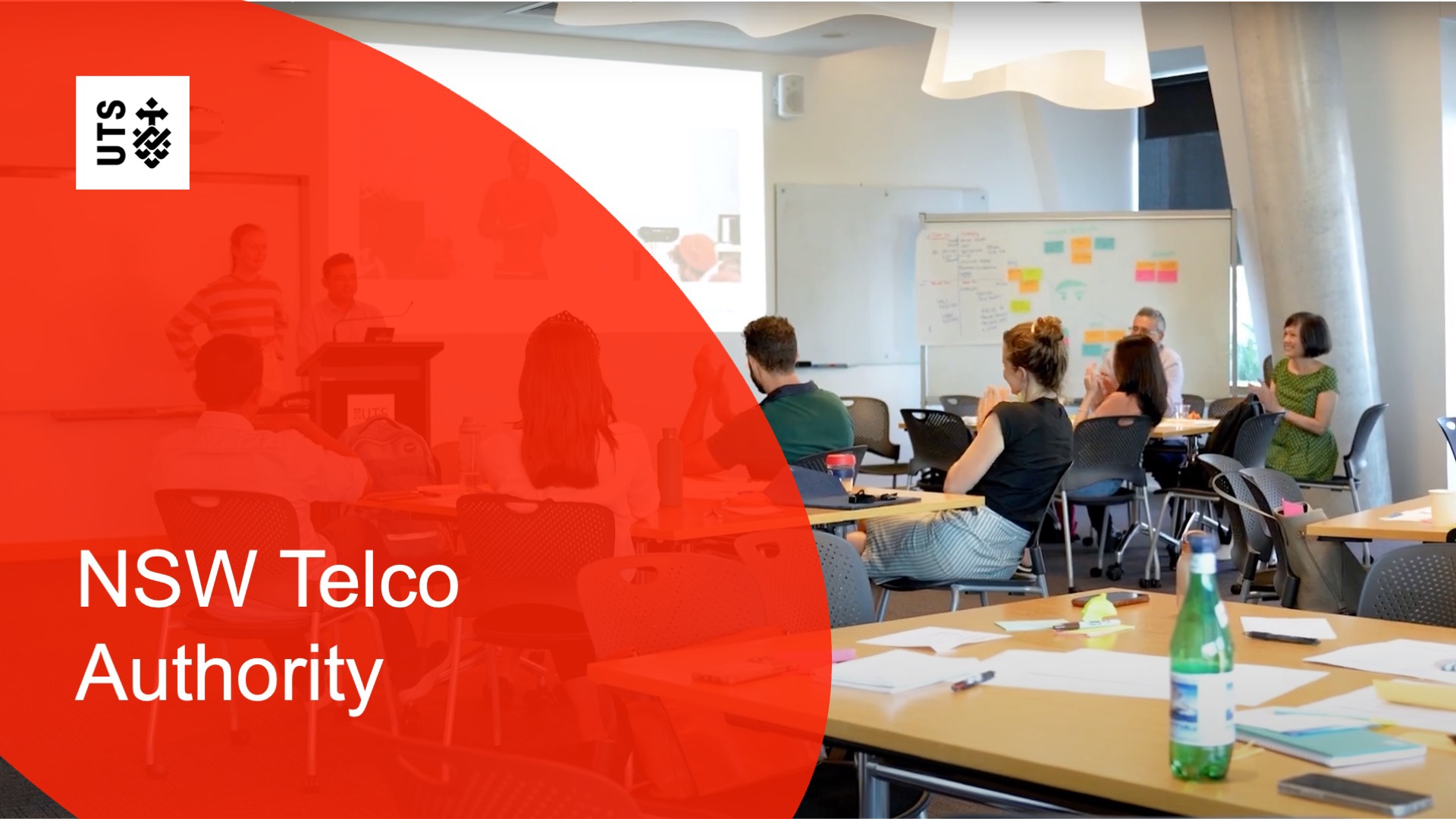 A classroom setting with people seated, engaging in a workshop. Papers and bottles on tables, a screen in the background. UTS logo and "NSW Telco Authority" text.