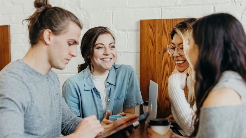 Four young adults sit together at a wooden table, engaged in conversation. They are smiling and using a tablet and a laptop, creating a friendly, collaborative atmosphere.