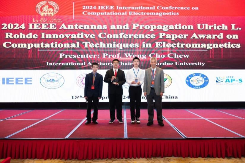Four individuals stand on a stage, proudly holding awards at the 2024 IEEE conference. A large banner in the background displays conference details.