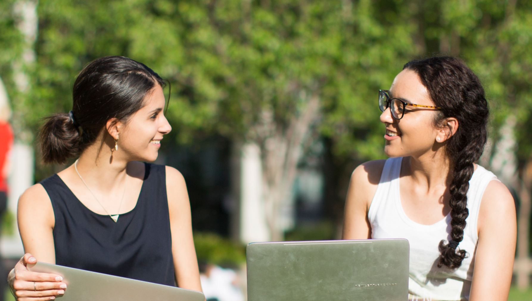 Two women sit outdoors with laptops, engaged in conversation. Both are smiling, creating a warm, friendly atmosphere. Trees are visible in the background.