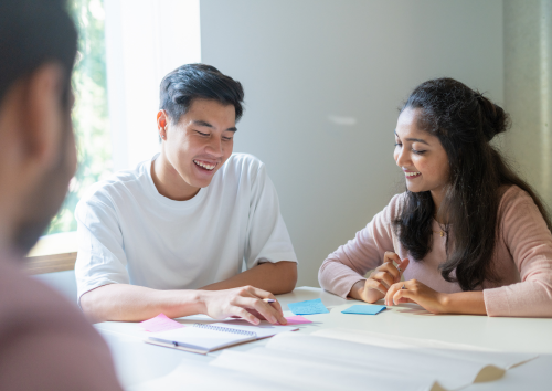 Two young adults sit at a table, smiling and discussing notes with sticky notes and a notebook, while another person is partially visible in the foreground.