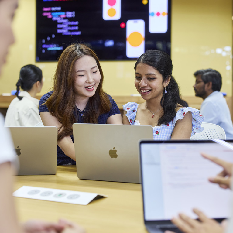 A group of young professionals collaborates at a table with laptops. Two women smile while discussing. A vibrant screen in the background displays visuals, creating an engaging workplace atmosphere.