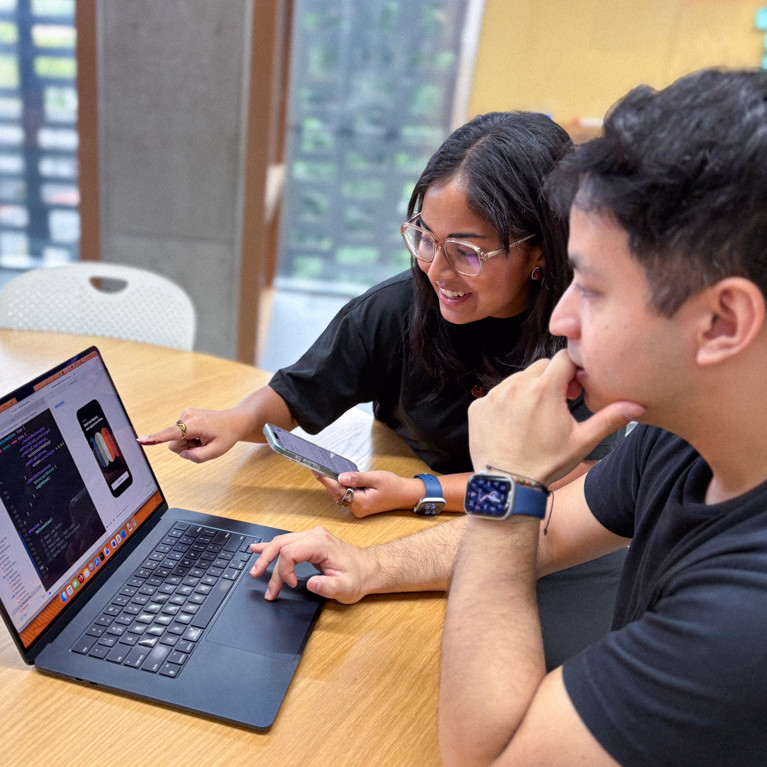 Two people collaborate at a table, reviewing code on a laptop, with an app design open. They appear focused and engaged in discussion.