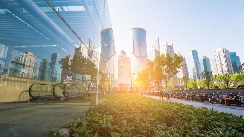 Modern cityscape with tall glass skyscrapers reflecting sunlight, lush green shrubs in the foreground, and a row of motorcycles on a sunlit street.