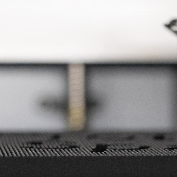 3D print head and black plastic part in focus showing material deposition and printing in process
