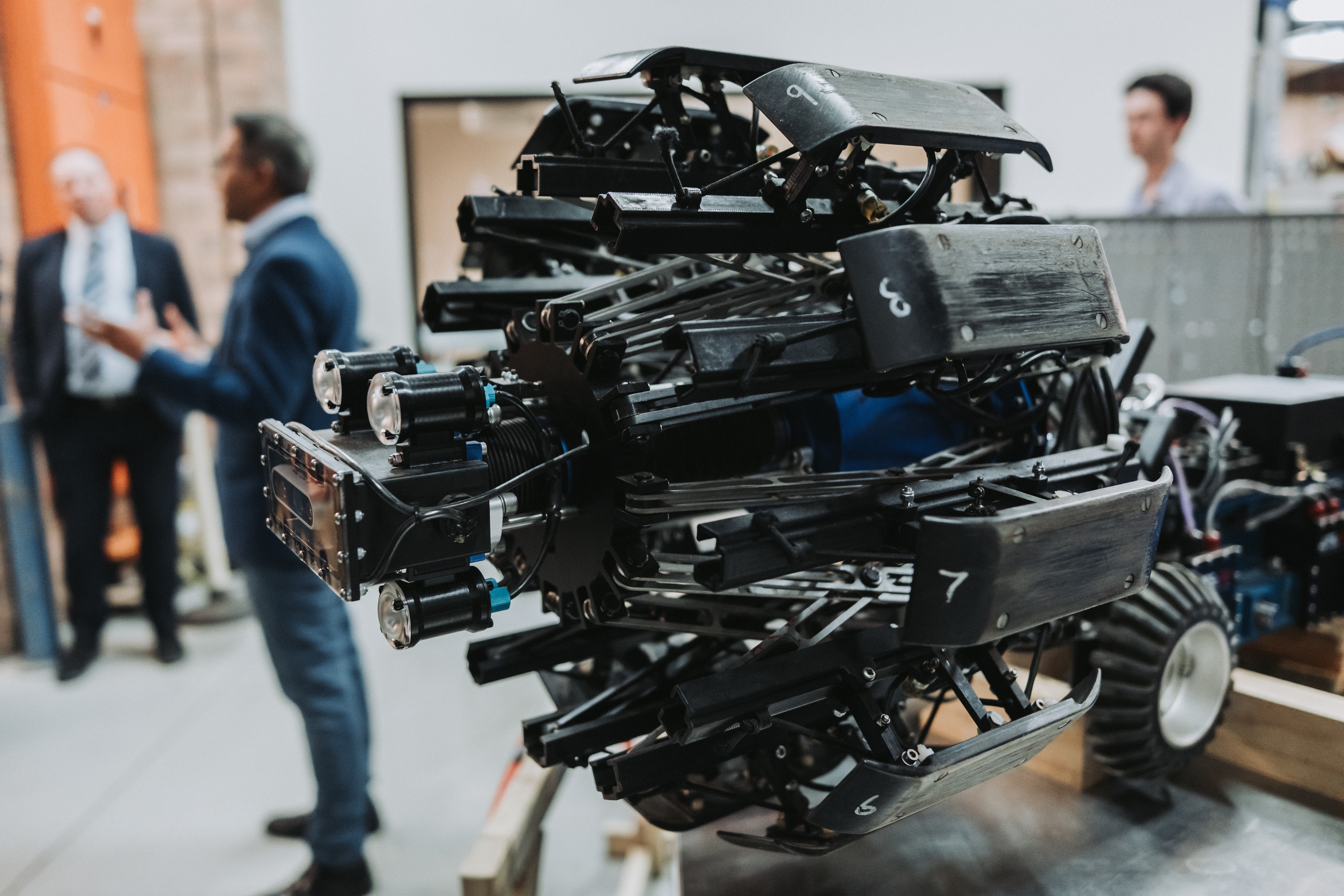A complex robotic mechanism with gears, rods, and wheels is in focus. Blurred background shows three people engaged in discussion, suggesting innovation.