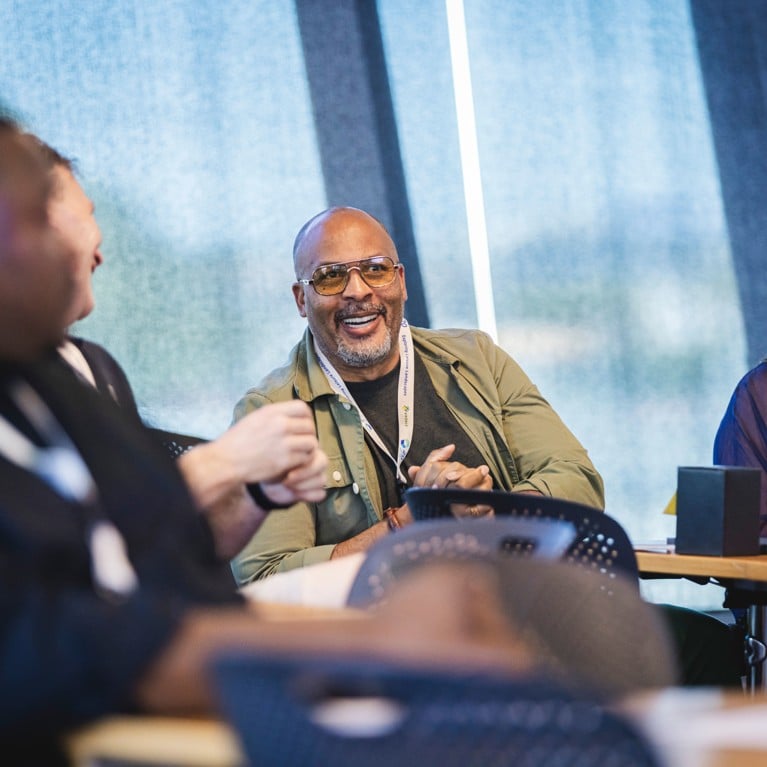 Participants seated around a meeting table during a collaborative workshop, with discussion taking place among attendees wearing conference lanyards.